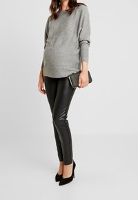 Kobieta w ciąży ubrana w szary prążkowany sweter, czarne legginsy o skórzanym wyglądzie, czarne szpilki i trzymająca czarną kopertówkę.