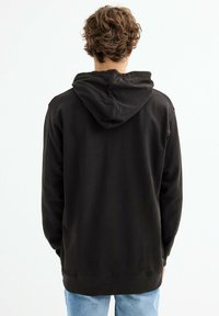 Sudadera con capucha oscura y cordón, textura suave, puños y dobladillo acanalados. Llevada sobre unos jeans de color azul claro, vista desde atrás.