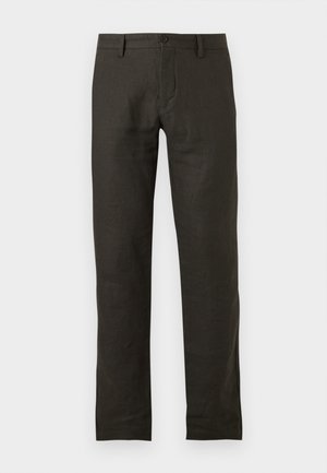 Homme debout, les mains dans les poches, portant une chemise jaune clair à manches courtes boutonnée, un pantalon foncé, des sandales noires et des lunettes de soleil.