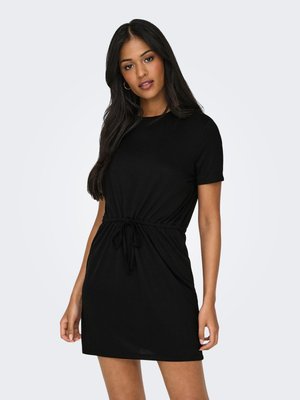 Jonge vrouw met lang donker haar, gekleed in een korte zwarte jurk met een strakke taille en korte mouwen, staand tegen een eenvoudige achtergrond.