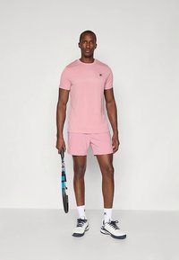 Ανδρικό αθλητικό σετ που περιλαμβάνει ένα εφαρμοστό, κοντομάνικο ροζ t-shirt και ένα ταιριαστό ροζ σορτσάκι. Κρατάει μια ρακέτα τένις και φοράει λευκά αθλητικά παπούτσια.