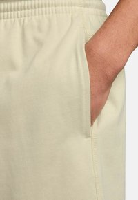 Beige jogger-housut, jotka on valmistettu pehmeästä, joustavasta kankaasta. Niissä on elastinen vyötärö ja sivutasku. Saumojen pitkin näkyvää ompelua.