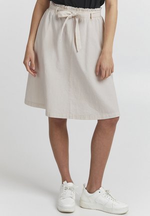 Jupe midi en coton beige avec une taille élastique froncée, une ceinture à nouer en tissu et des poches latérales. Portée avec des baskets blanches.