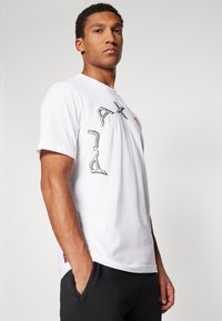 White cotton t-shirt featuring printed black graphics on the front and a small red logo on the side seam. Short sleeves, relaxed fit.