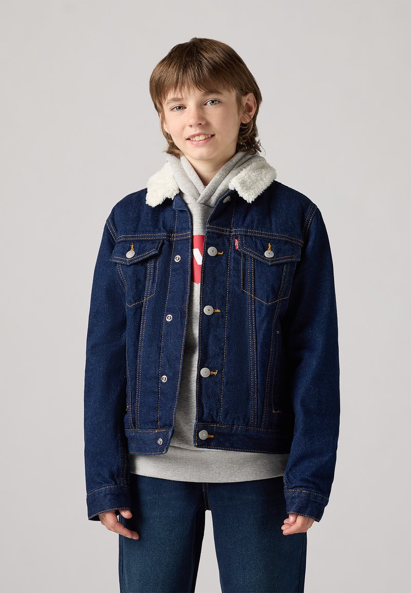 Giacca di jeans di colore blu scuro, con colletto in sherpa, accessori in metallo argentato e due tasche sul petto con dettagli di cucitura visibili.