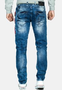 Jeans in denim blu con una finitura leggermente sbiadita, cinque tasche e un dettaglio con cerniera rossa in contrasto sul lato. Accentuati da sneakers bianche.