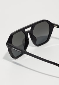 Ochelari de soare negri cu rame din plastic unghiulare și lentile gri. Au detalii din metal argintiu și marca "MICHAEL KORS" pe tijă.