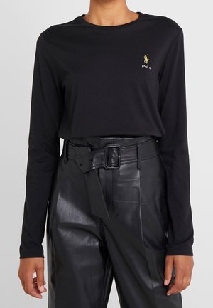 Osoba w czarnej koszulce Polo z długim rękawem z złotym logo, wpuszczona w wysokowciskane czarne skórzane spodnie z szerokim paskiem i kwadratową klamrą.