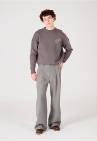 Sweatshirt en tricot gris avec col ras du cou et emblème, associé à un pantalon gris taille haute plissé et des chaussures marron, sur fond uni.