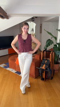 Jeune femme en haut sans manches violet et pantalon blanc, debout les mains sur les hanches dans un salon lumineux avec un rameur et une grande plante.