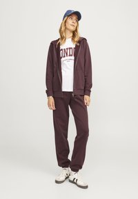 Felpa con zip color bordeaux e pantaloni della tuta abbinati, t-shirt bianca con stampa "LONDON", berretto blu e sneakers bianche con accenti neri.