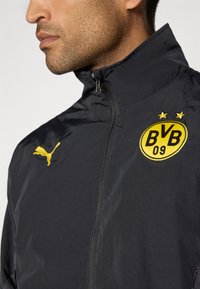 Schwarze Zip-Jacke mit hohem Kragen, die ein gelbes Puma-Logo und ein rundes schwarz-gelbes BVB 09-Emblem auf der Brust zeigt.