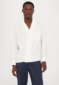 Cămașă albă cu mânecă lungă, confecționată dintr-un material ușor, cu guler cu tăietură, buzunar frontal și închidere cu nasturi. Purtați-o cu pantaloni bleumarin.