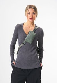 Olivgrüne Crossbody-Slingtasche mit strukturierter Oberfläche, verstellbarem Riemen und Reißverschlusstasche, getragen über einem grauen gerippten Oberteil.