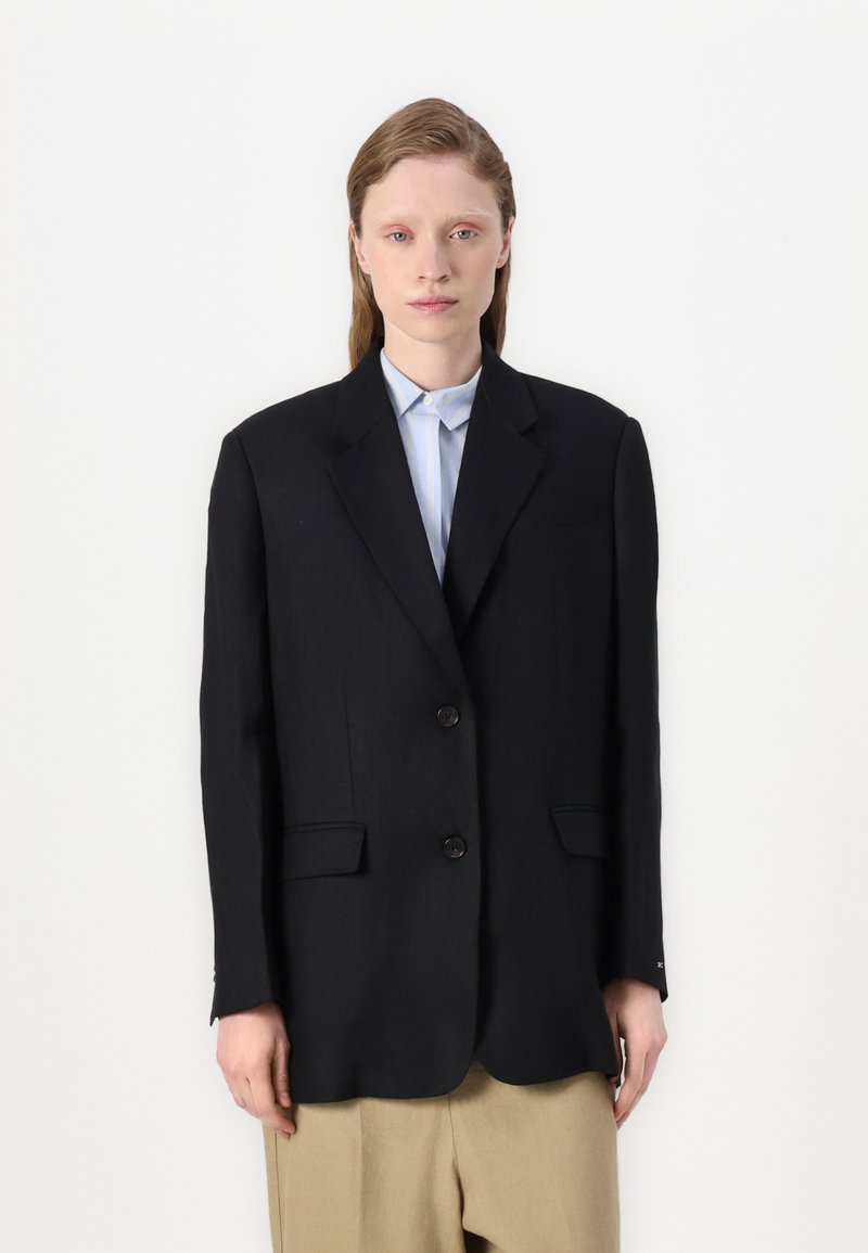 Blazer noir avec deux boutons, revers crantés et poches latérales. Porté par-dessus une chemise à col bleu clair et un pantalon beige.