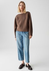 Maglione marrone, con texture, dalla forma squadrata e scollatura ampia, abbinato a jeans azzurri chiari, dalla vestibilità comoda, con tasche frontali e orlo sfrangiato. Scarpe scure.