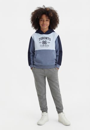 Lokkis juustega laps kannab sinist ja tumesinist Toronto kapuutsi, halli dressipükse ja valgeid kõrge säärega tossusid, seisab kätega taskutes.