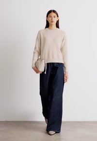 Licht beige wollen trui met een gestructureerde afwerking, gecombineerd met oversized donkere spijkerbroeken. Model houdt een zachte, ronde crèmekleurige handtas vast.