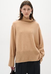 Pulover cu guler înalt de tip camel, realizat dintr-un material tricotat moale, cu o croială lejeră, manșete tale ribonate și un design minimalist. Pantaloni negri și o geantă.