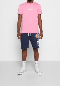 Růžové bavlněné tričko s kulatým výstřihem a bílým logem "ellesse"; námořnické šortky s bílým logem a oranžovo-bílým akcentem. Bílé sneakers s vysokým střihem.