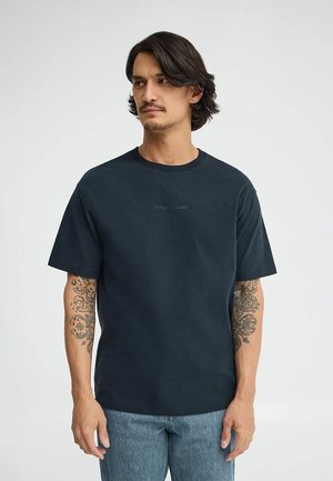 Mann mit schulterlangem Haar, der ein schwarzes "Polo Club"-T-Shirt und blaue Jeans trägt, Tattoos auf beiden Unterarmen zeigt und zur Seite blickt.
