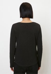 Person with straight black bob haircut wearing a plain long-sleeve black top and black pants, facing a white background.