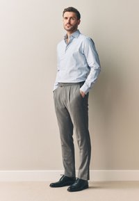 Uomo in piedi con le mani in tasca, indossa una camicia azzurra, pantaloni grigi e scarpe eleganti nere, davanti a un muro beige.