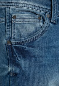 Vaqueros de mezclilla azul claro con una textura desgastada, que cuentan con un bolsillo para monedas, remaches de metal y costuras amarillas contrastantes.