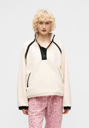 Göndör szőke hajú személy krémszínű polár pulóvert visel fekete szegéllyel és rózsaszín mintás nadrágot, egyszerű háttér előtt áll.