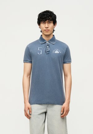 Joven con una camisa polo azul desgastada de Hollister con logo y número 5, combinada con vaqueros de mezclilla claros, de pie frente a un fondo blanco.