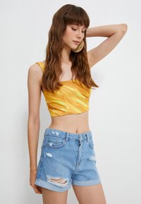 Joven con cabello largo y ondulado, lleva pendientes grandes y redondos, un top corto amarillo y shorts de denim desgastados de color azul claro, posando frente a un fondo blanco.