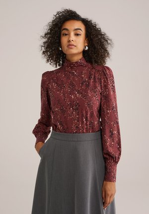 Une femme aux cheveux bouclés porte un haut à motif bordeaux et une jupe grise, posant les mains dans les poches, sur un fond neutre.