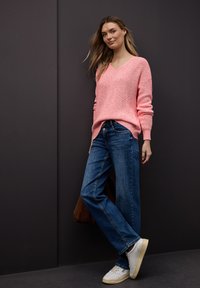 Femme appuyée contre un mur sombre, portant un pull en tricot rose, un jean bleu, des baskets blanches et tenant un sac marron.