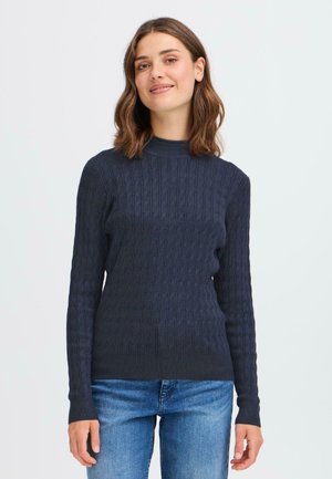 Mørkeblå teksturert genser med høy hals og ribbestrikkede mansjetter, med et kabelstrikket mønster, kombinert med lyseblå jeans.