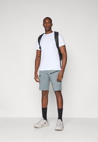T-shirt blanc à manches courtes avec logo, short cargo gris clair avec poches zippées, sac à dos noir, chaussettes noires et baskets légères.