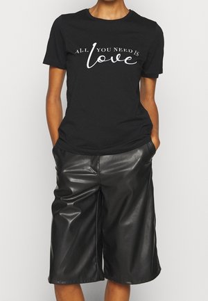 Persoon draagt een zwart T-shirt met de tekst "All you need is love" en zwarte nepleren korte broek tot op de knie, handen in de zakken, staand tegen een witte achtergrond.