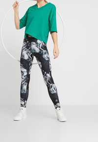 Zielona bluzka z krótkim rękawem i dekoltem w serek, zestawiona z czarnymi legginsami w abstrakcyjne białe wzory. Modelka nosi białe sneakersy.