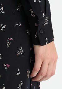 Svart tyg med små blommönster i rosa, vitt och grönt. Knappdetalj vid ärmslutet nära handleden, slät textur.