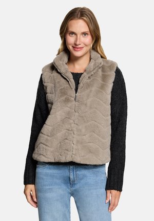 Woman wearing light brown fuzzy zip-up vest over black sweater and blue jeans, smiling at the camera against a plain background.