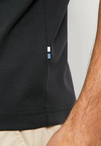 Schwarzes Poloshirt aus strukturiertem Stoff, mit einem kleinen, mehrfarbigen Logo an der Seitennaht und einer körperbetonten Passform.