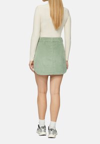 Mini-jupe en velours côtelé vert avec une taille ajustée et une texture striée subtile, associée à des chaussures de sport argentées et des chaussettes blanches mi-mollet.