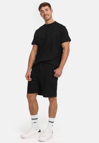 Ung mand, der smiler, iført en sort T-shirt og shorts, hvide sokker med sorte striber og hvide sneakers, stående foran en ensartet hvid baggrund.