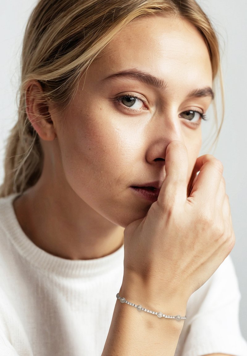 Bracciale d'argento con piccole perline rotonde e pietre trasparenti, indossato su un polso, su uno sfondo chiaro, mano che tocca delicatamente il viso.