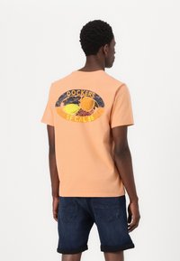 Tricou din bumbac de culoare piersică, cu un imprimeu grafic pe spate. Designul include fructe și text în culorile albastru și galben, are mâneci scurte și o croială clasică.
