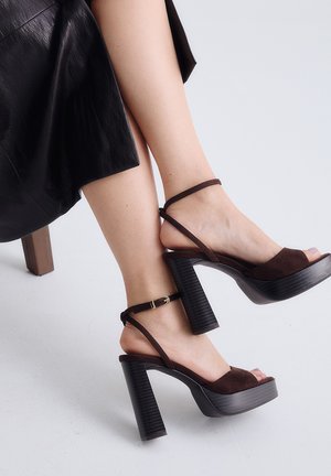 Piernas cruzadas con sandalias de plataforma de tacón alto de color marrón oscuro con tiras en el tobillo, combinadas con una falda negra de cuero.