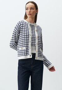 Marineblauwe en witte geblokte cardigan met ronde crèmekleurige knopen, geribde manchetten en twee voorzakken. Ggedragen over een lichte trui en donkere spijkerbroek.