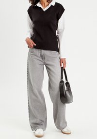 Gilet noir sans manches en maille porté sur une chemise blanche à col, associé à un jean large gris clair et des baskets blanches, tenant un sac noir.