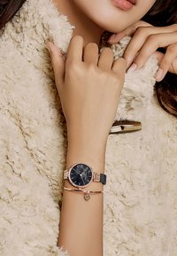 Montre noire avec des accents en rose gold et un cadran à motif, portée avec un bracelet en rose gold présentant un détail de logo, sur un fond crème texturé.