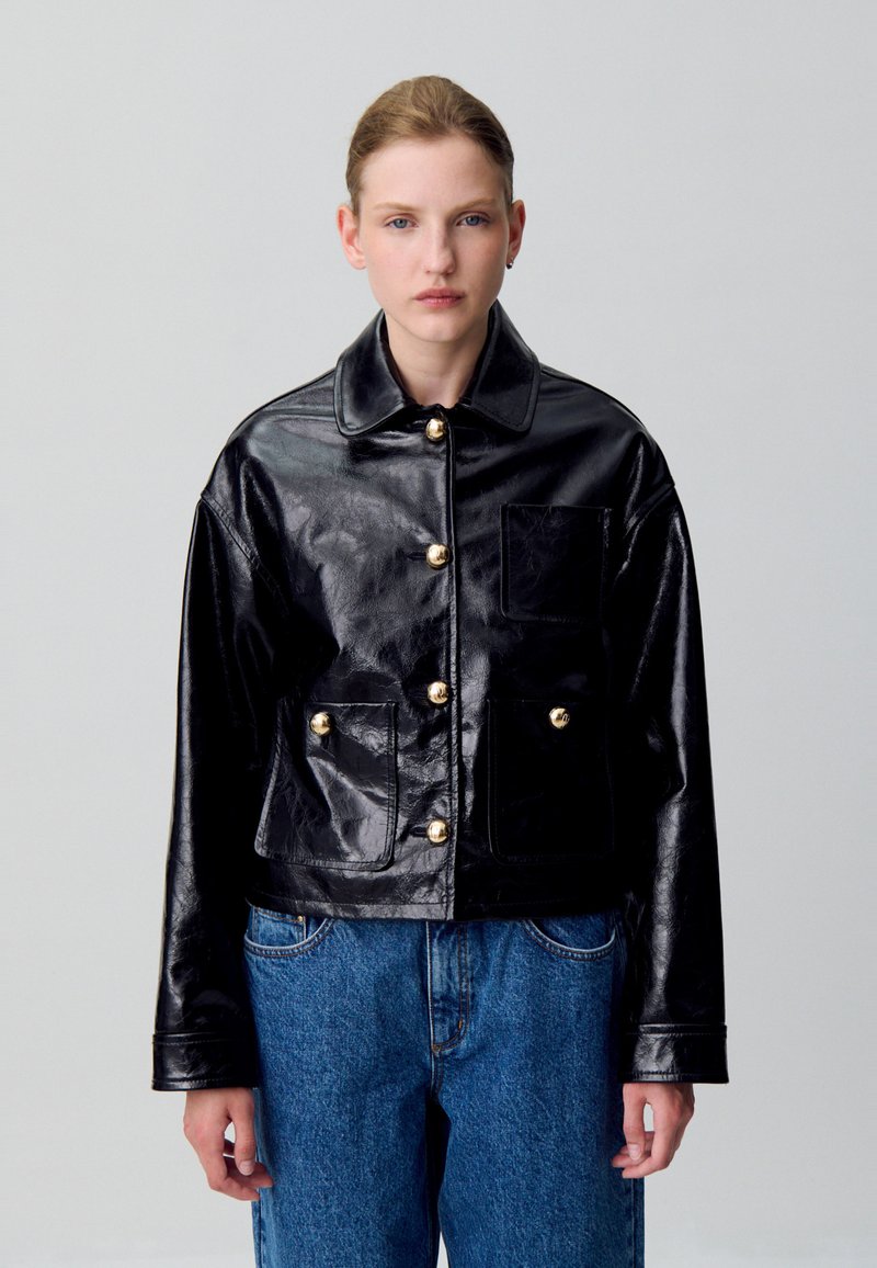 Giacca croppata in pelle nera con una texture lucida, dotata di chiusura con bottoni, bottoni dorati e due tasche frontali.