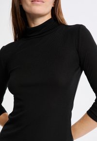 Donna che indossa un maglione aderente nero a maniche lunghe con le mani sui fianchi, sfondo bianco.